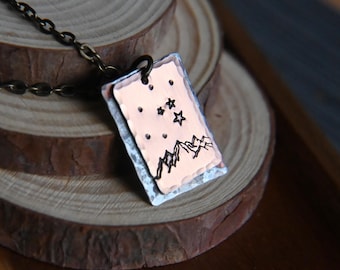 Könnte beinhalten: Eine kupferfarbene Anhängerkette mit einem rechteckigen Anhänger mit einem Berg- und Sterndesign. Der Anhänger hängt an einer dunklen Kette. Die Halskette wird auf einer Holzoberfläche präsentiert.