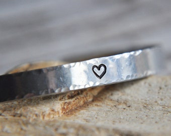 Könnte beinhalten: Ein silbernes, gehämmertes Armband mit einem kleinen Herz-Design.