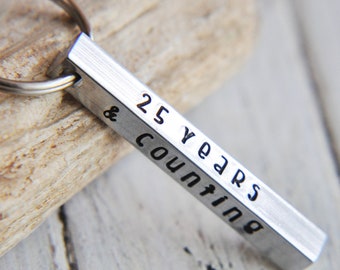 May include: Silver keychain with a bar charm engraved with the text "25 years & counting".