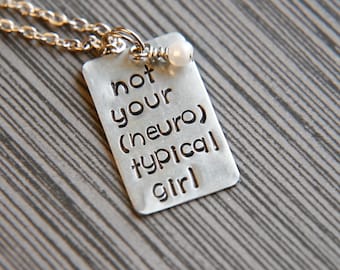 Könnte beinhalten: Silberne Halskette mit einem rechteckigen Anhänger, auf dem "not your (neuro) typical girl" steht, und einer kleinen weißen Perle.