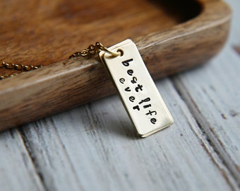 Könnte beinhalten: Goldkette mit einem rechteckigen Anhänger, auf dem "best life ever" steht.