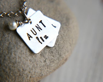 Puede incluir: Un collar de plata con un dije que dice "AUNT" y una pequeña perla. El dije está unido a una cadena de plata.