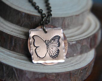 Könnte beinhalten: Kupferfarbene Halskette mit Anhänger, die ein Schmetterlingsdesign zeigt. Der Schmetterling ist schwarz auf einem quadratischen Anhänger mit gehammerter Textur geätzt. Die Kette ist dunkelbraun, vor einem Holz Hintergrund.