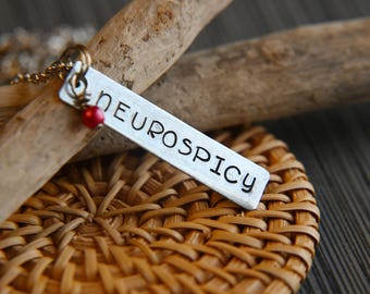 Könnte beinhalten: Eine silberne Halskette mit einem rechteckigen Anhänger, auf dem "Neurospicy" steht. Der Anhänger hängt an einer Kette mit einer kleinen roten Perle.