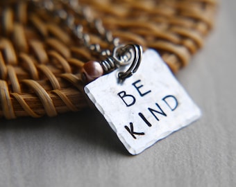 Könnte beinhalten: Eine silberne Halskette mit einem quadratischen Anhänger, auf dem in schwarzen Buchstaben "BE KIND" steht. Der Anhänger ist an einer silbernen Kette mit einer kleinen rosa Perle befestigt.