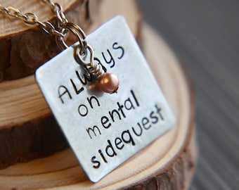 Könnte beinhalten: Silberne Halskette mit einem quadratischen Anhänger, auf dem "Always on a mental sidequest" steht. Der Anhänger hat eine kleine Kupferperle.