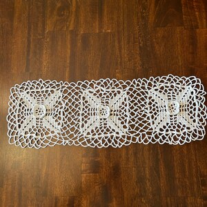 May include: White crocheted table runner with three square designs featuring a floral pattern in the center. The runner is on a dark brown wooden surface.
