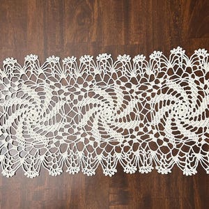 May include: Cream-colored, rectangular crochet table runner with a spiral design. The openwork pattern features a repeating floral edge. The runner is placed on a dark brown wooden surface.