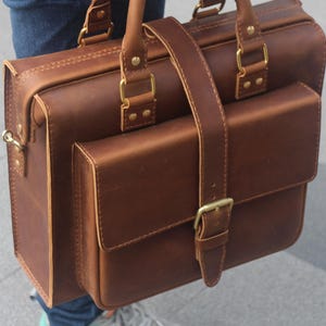 May include: A brown leather briefcase with a front pocket and a buckle closure. The bag has a strap and two handles.