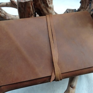 May include: A brown leather journal with a leather strap closure. The journal is lying on a piece of driftwood.