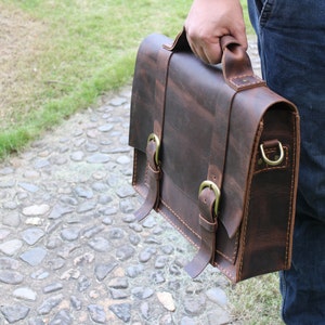 May include: A brown leather briefcase with brass buckles and a strap. The briefcase has a distressed look and is being held by a person.