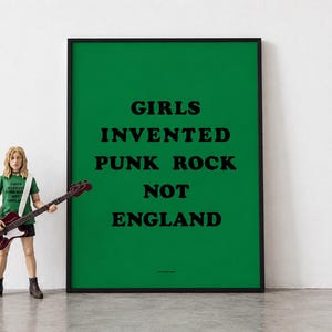 Puede incluir: Un póster rectangular con fondo verde y texto negro que dice GIRLS INVENTED PUNK ROCK NOT ENGLAND. Al lado hay una figura de acción rubia con una camiseta verde con el mismo texto, pantalones cortos negros y una guitarra bajo roja.