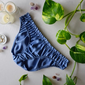 May include: A blue satin underwear with a gathered, ruffled design. The underwear is on a white surface with green leaves and small stones.