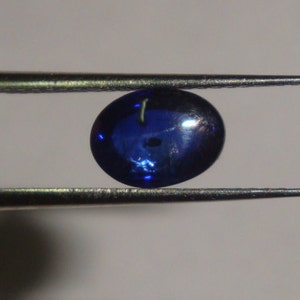 May include: A dark blue oval-shaped gemstone with a slight shimmer, held by tweezers.