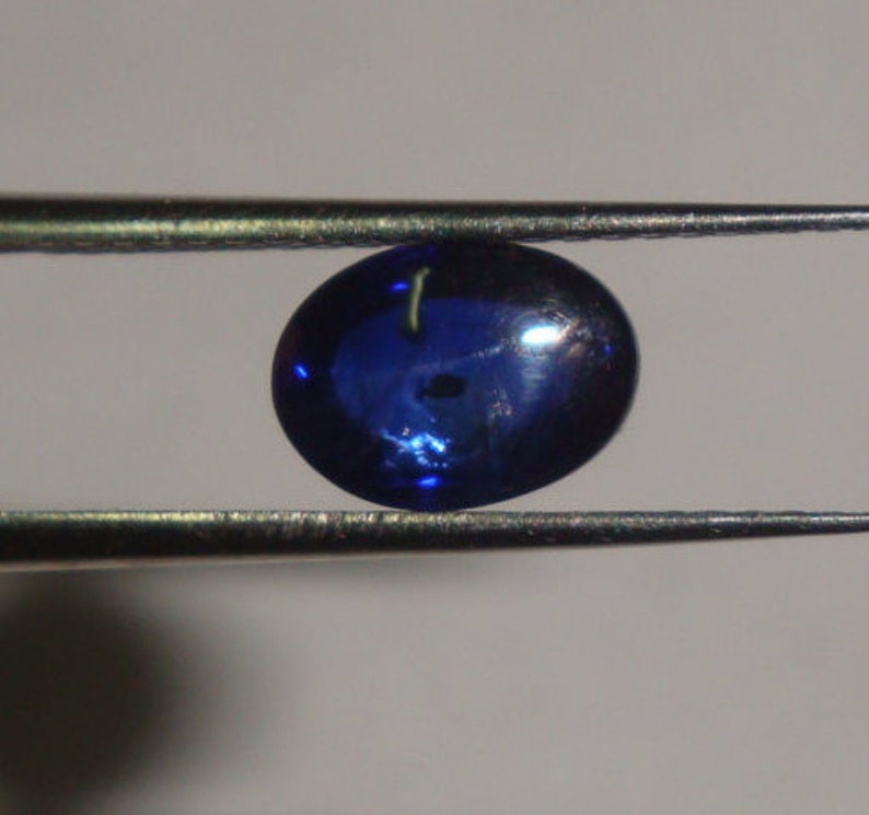 May include: A dark blue oval-shaped gemstone with a slight shimmer, held by tweezers.