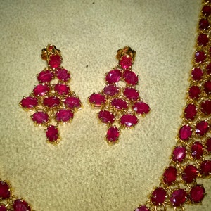May include: A gold necklace and earrings set with red ruby stones. The necklace is a long, thin chain with a pendant that is made up of several rows of ruby stones. The earrings are also made up of several rows of ruby stones.