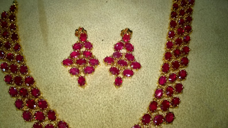 May include: A gold necklace and earrings set with red ruby stones. The necklace is a long, thin chain with a pendant that is made up of several rows of ruby stones. The earrings are also made up of several rows of ruby stones.
