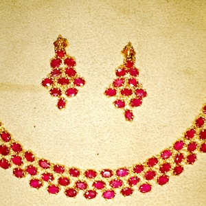 May include: A gold necklace and earrings set with red ruby stones. The necklace is a curved design with multiple rows of ruby stones. The earrings are dangle style with multiple ruby stones.