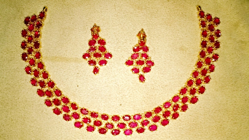 May include: A gold necklace and earrings set with red ruby stones. The necklace is a curved design with multiple rows of ruby stones. The earrings are dangle style with multiple ruby stones.