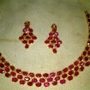May include: A gold necklace and earrings set with red ruby stones. The necklace has three rows of oval-shaped stones.