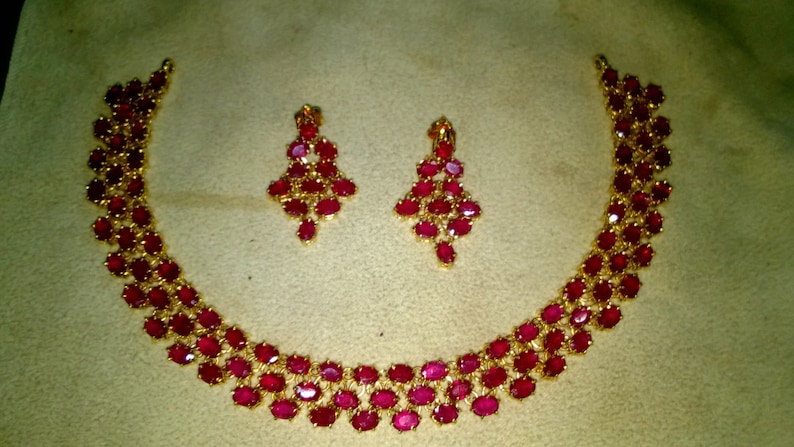 May include: A gold necklace and earrings set with red ruby stones. The necklace has three rows of oval-shaped stones.