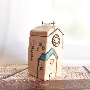 Puede incluir: Una caja de cerámica con forma de casa con un esmalte marrón claro y detalles azules y naranjas. La caja tiene un pequeño corazón en la parte delantera y una ventana redonda con un diseño de sol amarillo. La caja tiene una pequeña cuerda de alambre con una cadena de cuentas en la parte superior.