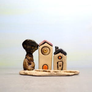 May include: A ceramic sculpture of three small houses with a tree in front. The houses are painted in shades of beige, pink, and black with orange and yellow details. The tree is brown and green.