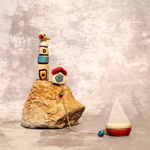 May include: A ceramic lighthouse figurine with a red roofed house and a blue flower on a rock base. A white sailboat with a red bottom is attached to the base with a gold chain.