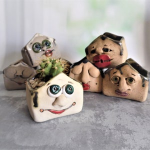 Op de afbeelding: Een set van zes keramische potten in de vorm van gezichten met verschillende uitdrukkingen. De potten zijn geschilderd in tinten bruin, beige en wit met groene en rode accenten. Een pot heeft een kleine cactusplant die erin groeit.