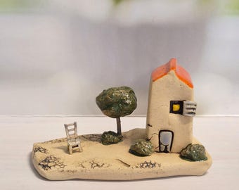 May include: A miniature ceramic house with an orange roof, a small tree, and a tiny chair on a tan base. The house has a yellow window and a black door. The scene evokes a quaint, rustic aesthetic.