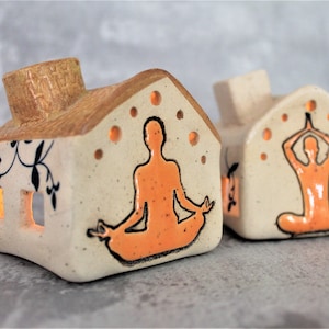 May include: Two ceramic house-shaped candle holders featuring orange yoga figures. One figure is in a seated meditation pose, the other is in a standing pose with arms raised.