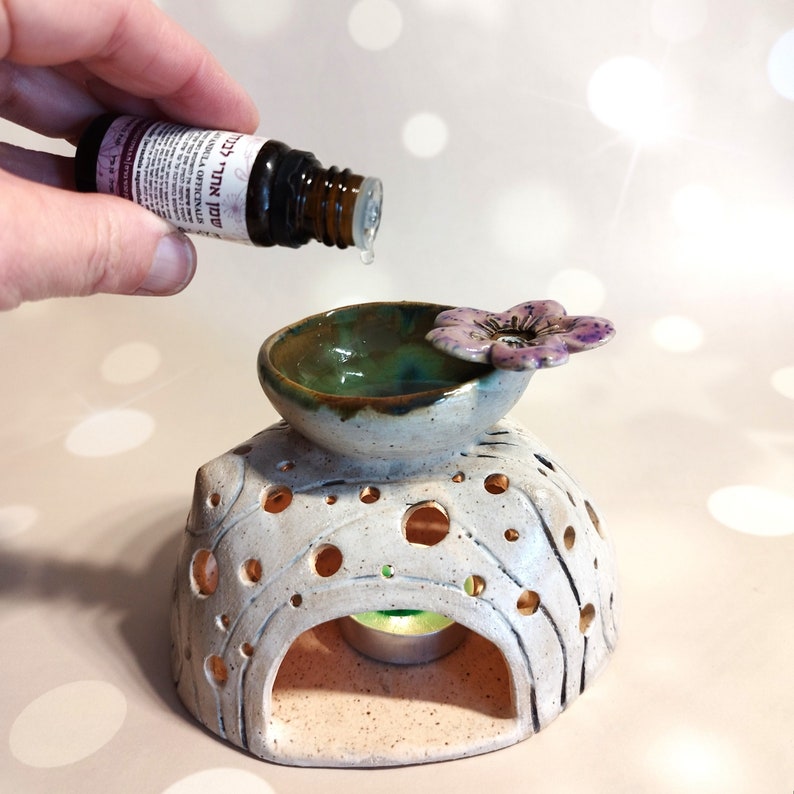 May include: White ceramic oil diffuser with a flower-shaped lid and a small bowl for essential oils. The diffuser has a hole in the centre for a tea light candle. A bottle of essential oil is being poured into the bowl.