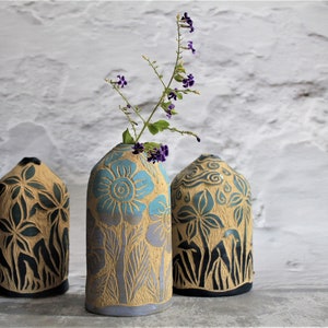Könnte beinhalten: Drei Keramikvasen mit Blumendesigns. Die Vasen sind in Braun-, Blau- und Schwarztönen gehalten. Die Vase in der Mitte hat ein hellblaues Blumendesign und hält einen Zweig mit violetten Blüten.