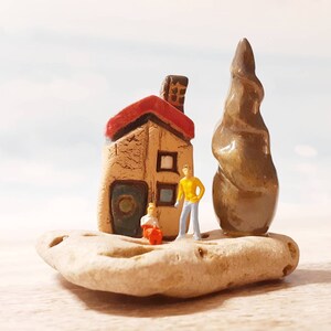 May include: A miniature ceramic house with a red roof, a brown tree, and two tiny figurines standing in front of it. The scene is set on a light brown rock.