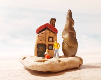 May include: A miniature ceramic house with a red roof, a brown tree, and two tiny figurines standing in front of it. The scene is set on a light brown rock.