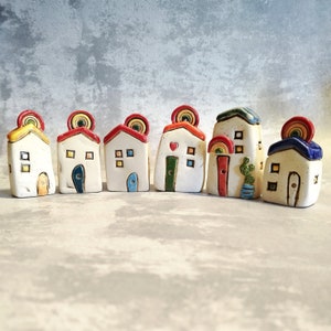 Może przedstawiać: Zestaw siedmiu małych, ręcznie malowanych domków ceramicznych z akcentami tęczy. Każdy domek ma unikalny projekt i kolorystykę, w tym czerwony, niebieski, zielony, żółty i biały.