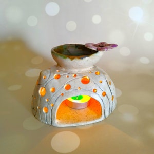 May include: White ceramic oil diffuser with a flower-shaped top and a small candle inside. The diffuser has a textured surface with holes and a round opening at the front.