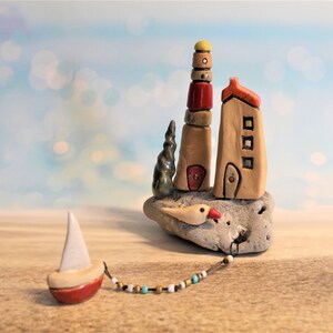 May include: A ceramic sculpture of a lighthouse, a house, and a small sailboat on a rock base. The lighthouse is multi-colored with a yellow top and red accents. The house is tan with a red roof and windows. The sailboat is red with a white sail. The sculpture is on a wooden surface.