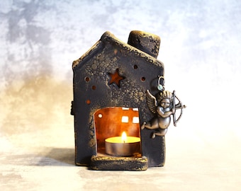 Könnte beinhalten: Ein kerzenhalter aus Keramik in Form eines Hauses mit goldener und schwarzer Oberfläche. Der Halter hat ein kleines Fenster mit einem brennenden Teelicht darin und eine Cherubfigur mit Pfeil und Bogen an der Seite.
