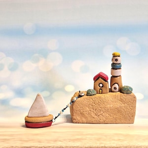 May include: A miniature ceramic seaside scene featuring a lighthouse, a small house, and a sailboat. The lighthouse is stacked with colorful rings, and the house has a red roof. The sailboat is red and white, connected to the scene by a beaded string.