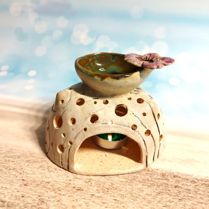May include: A ceramic oil burner with a flower design on the top. The burner has a small bowl for oil and a space for a tea light. The burner is white with brown and green accents.
