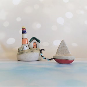 May include: A ceramic lighthouse with a white base and colorful stripes, a small house with a green roof and a red door, and a sailboat with a red bottom are connected by a string of blue beads. The scene is set on a white background with a blurred bokeh effect.