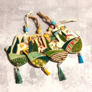 May include: Four ceramic ornaments with a whimsical village scene, each featuring a different design with green, yellow, and red accents. The ornaments are attached to strings with tassels.
