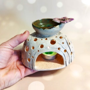 May include: White ceramic oil burner with a flower design on top and a small opening for a tea light. The burner has a textured surface with small holes.