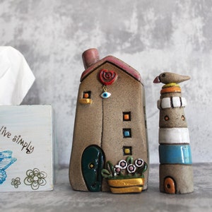 May include: Two handmade ceramic figurines, a house with a red heart and blue eye, and a lighthouse with a bird on top. The house has a green door and a flower pot with pink flowers. The lighthouse has white and blue stripes. Both figurines are on a gray background. The text "Live Simply" is on a blue and white box in the background.