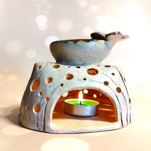 May include: A white ceramic oil burner with a small, round, green tea light candle inside. The burner has a decorative top with a flower design and a small bowl for essential oils.