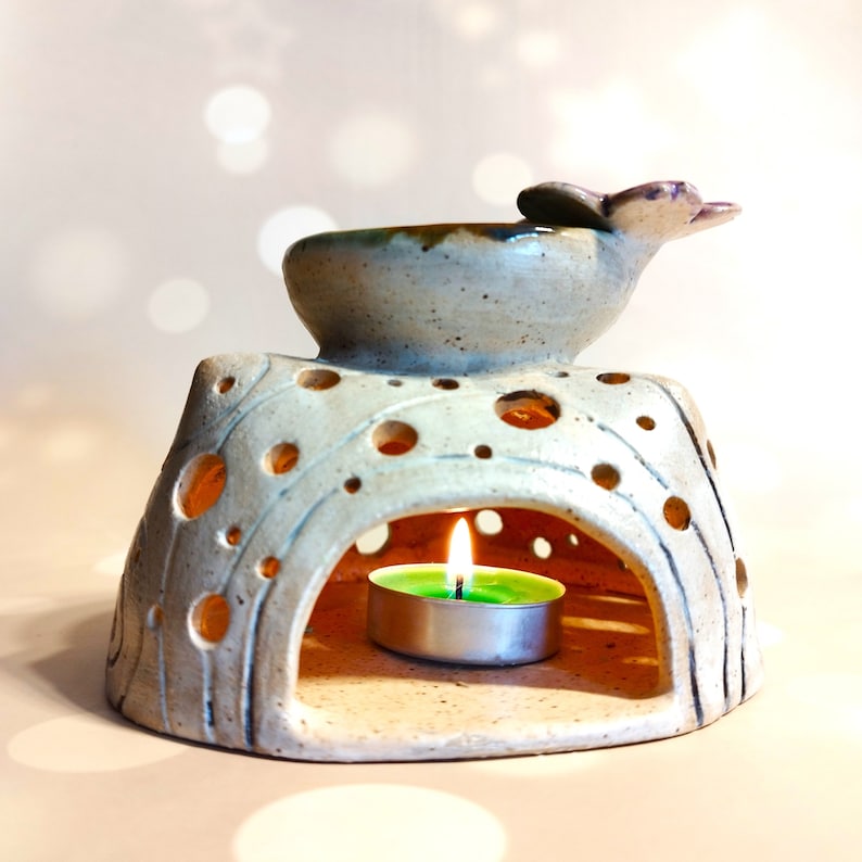 May include: A white ceramic oil burner with a small, round, green tea light candle inside. The burner has a decorative top with a flower design and a small bowl for essential oils.