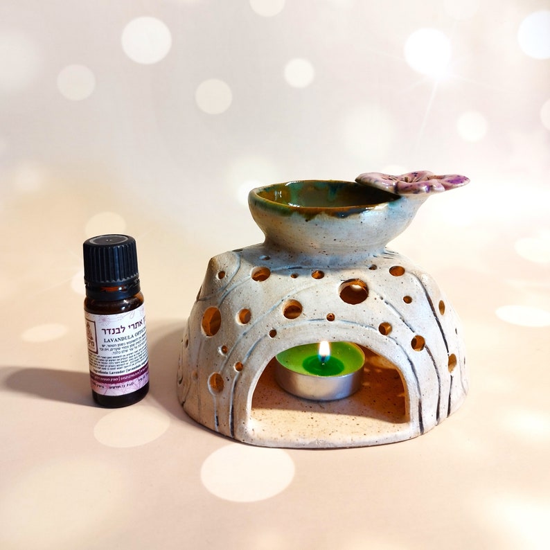 May include: A white ceramic essential oil diffuser with a green and brown top and a small flower detail. A small bottle of lavender essential oil is in the foreground. The diffuser has a small opening for a tea light candle.