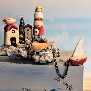 May include: Ceramic tissue box cover with a nautical theme. The cover features a small sailboat, a bird, a lighthouse, and several houses on a rocky base. The text "live simply" is written on the front of the box.