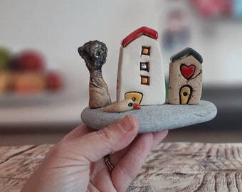 Ceramic sculpture of a whimsical village with miniature clay houses, a tree and a bird, on a natural beach stone